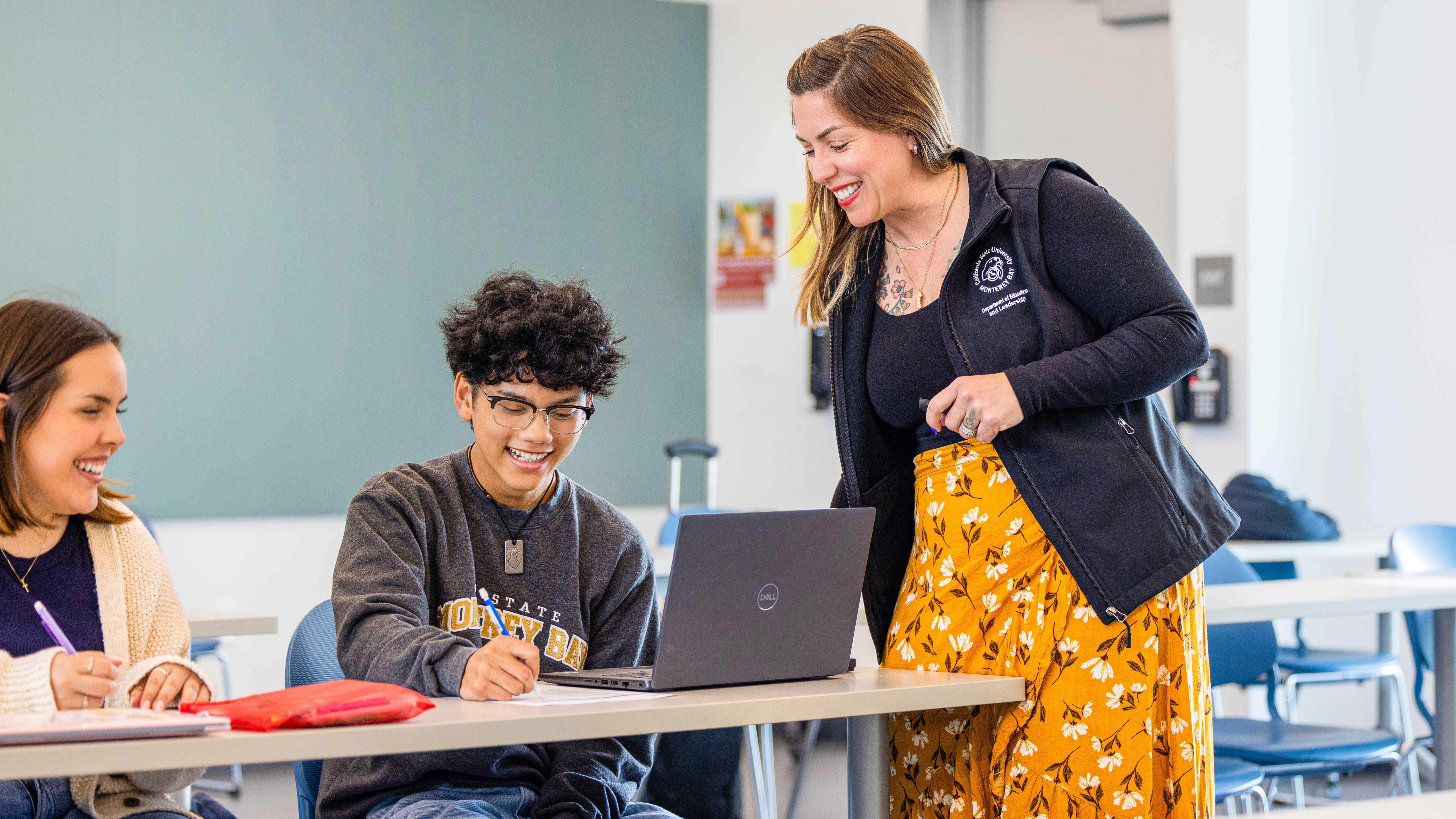 Professor smiling at two students on laptops in a class