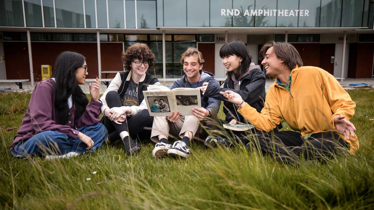 Five students sitting outside on the lawn looking at a book